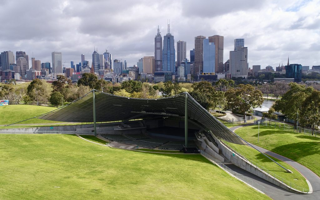 Aerial view of an open-air amphitheater with a distinctive curved canopy facing a city skyline.