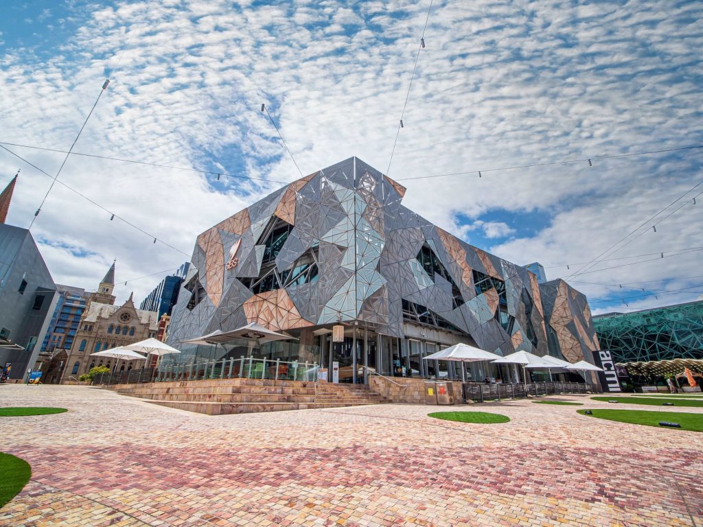A geometric, multi-faceted building at Federation Square under a blue sky.