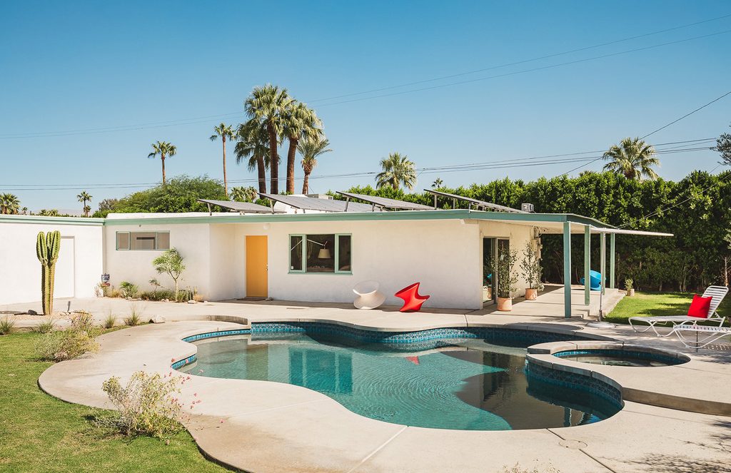 White mid-century modern single-story house with solar panels, a backyard swimming pool, and palm trees.