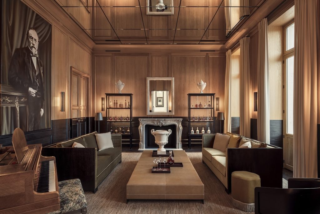 Warmly lit, elegant lounge lined with light wood panelling, dark green velvet sofas, a large central ottoman coffee table, and a classic upright piano, best interior designers in Paris