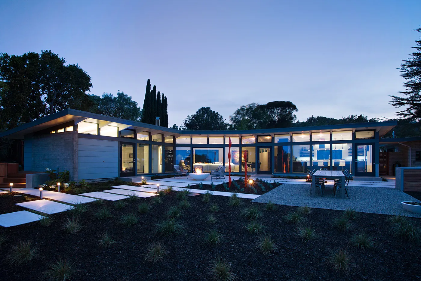 Exterior view at twilight of a modern L-shaped home with large glass windows and an illuminated patio.