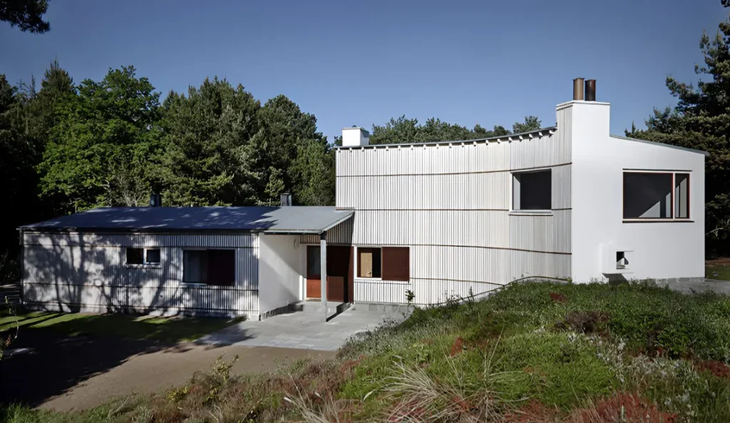 Modern white two-story summer house with a curved, textured facade set on a grassy hill surrounded by trees.