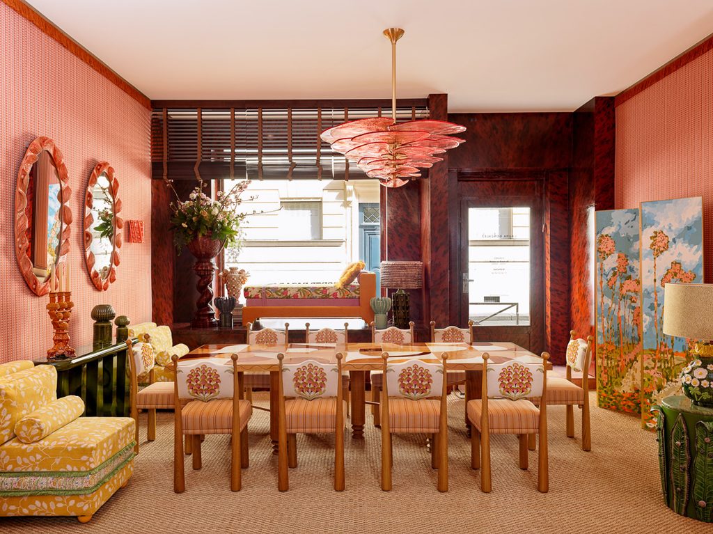 Eclectic dining room styled with salmon pink textured walls, a floral-upholstered dining set, a red petal pendant lamp, and vibrant yellow armchairs, best interior designers in Paris
