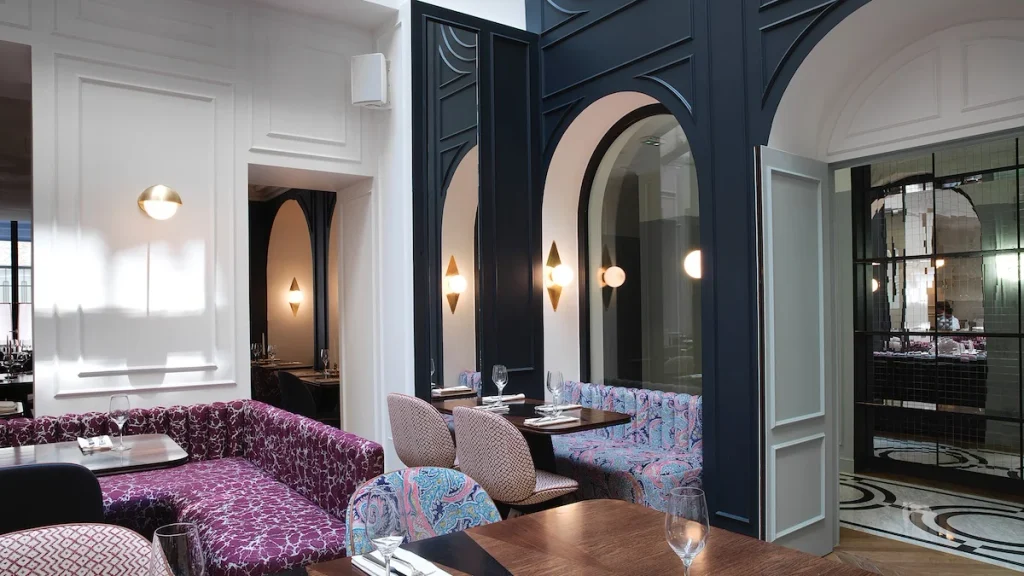 Elegant dining area with high white walls, navy archways, patterned purple and pink booth seating, and subtle wall sconce lighting, best interior designers in Paris