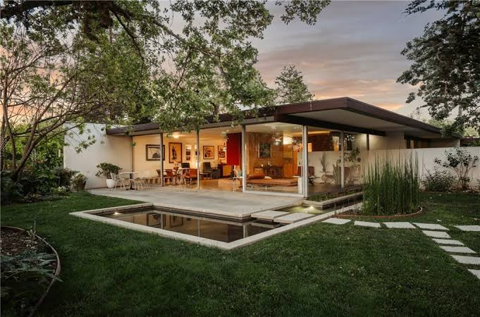 Single-story modern house at dusk with open glass sliding doors leading to a patio and a small garden pond.