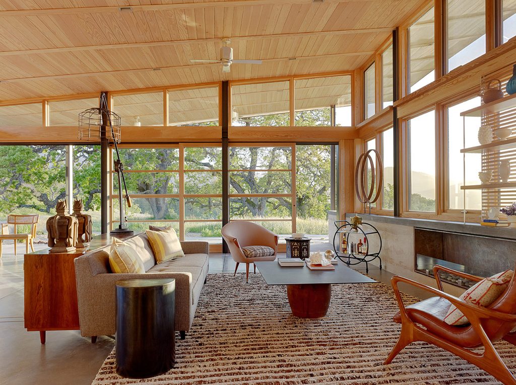 Sunlit living room with floor-to-ceiling glass doors, wood paneling, a beige sofa, and views of oak trees.