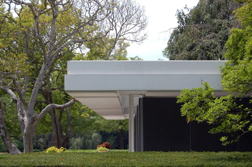 Exterior detail of a sleek modern white roof structure supported by a pillar, surrounded by lush trees.