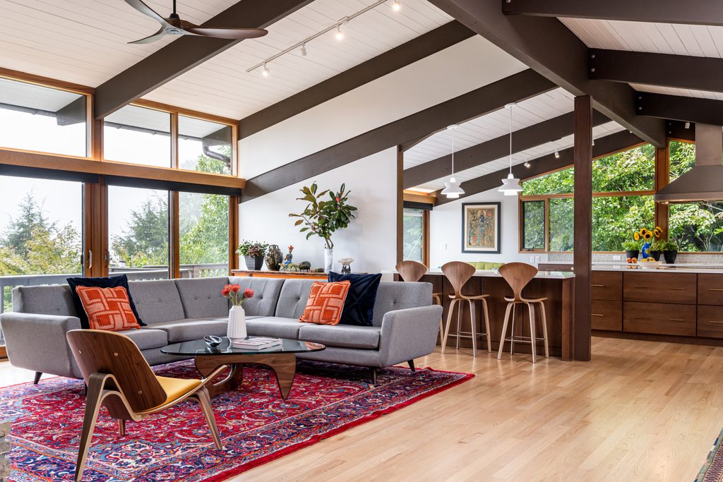 Mid-century modern living room featuring exposed dark wood ceiling beams, a grey sectional sofa, and a red patterned rug