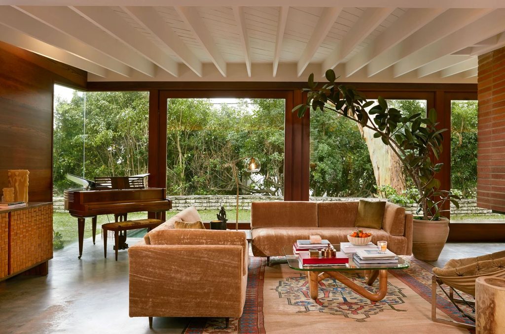 Mid-century interior with brown velvet sofas, floor-to-ceiling glass walls, a grand piano, and a large indoor plant.