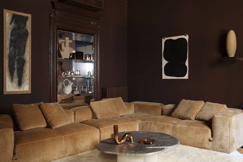 Moody lounge interior with dark brown walls, a plush tan modular sectional sofa, and a natural stone coffee table. top interior design firms in milan