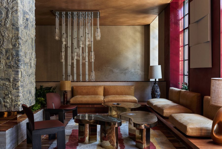Luxurious lounge interior featuring brass coffee tables, tan leather seating, rich red wall paneling, and an elaborate hanging glass chandelier. top interior design firms in milan