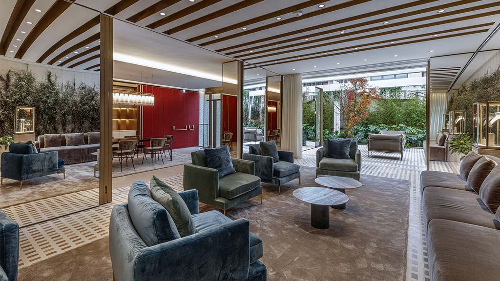Modern open-plan seating hall with slatted ceiling details, plush blue armchairs, and large glass walls looking out to greenery. top interior design firms in milan