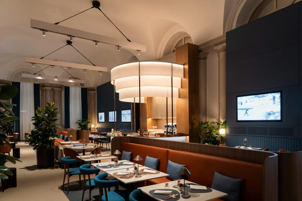Spacious restaurant and lounge area featuring terraced ceiling lights, blue accent chairs, and warm leather booth seating. top interior design firms in milan