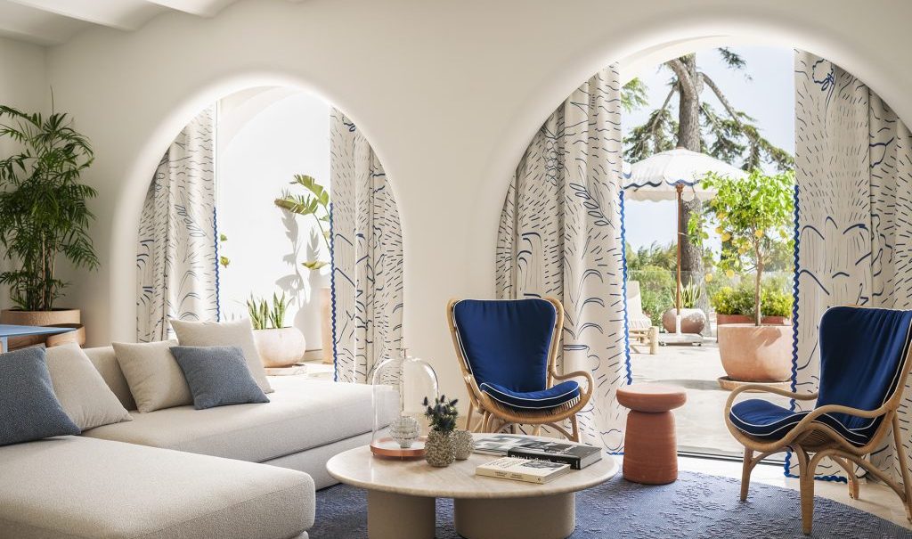 Bright Mediterranean-style lounge opening through arched doorways to a sunlit terrace, decorated with blue accents and patterned curtains. top interior design firms in milan