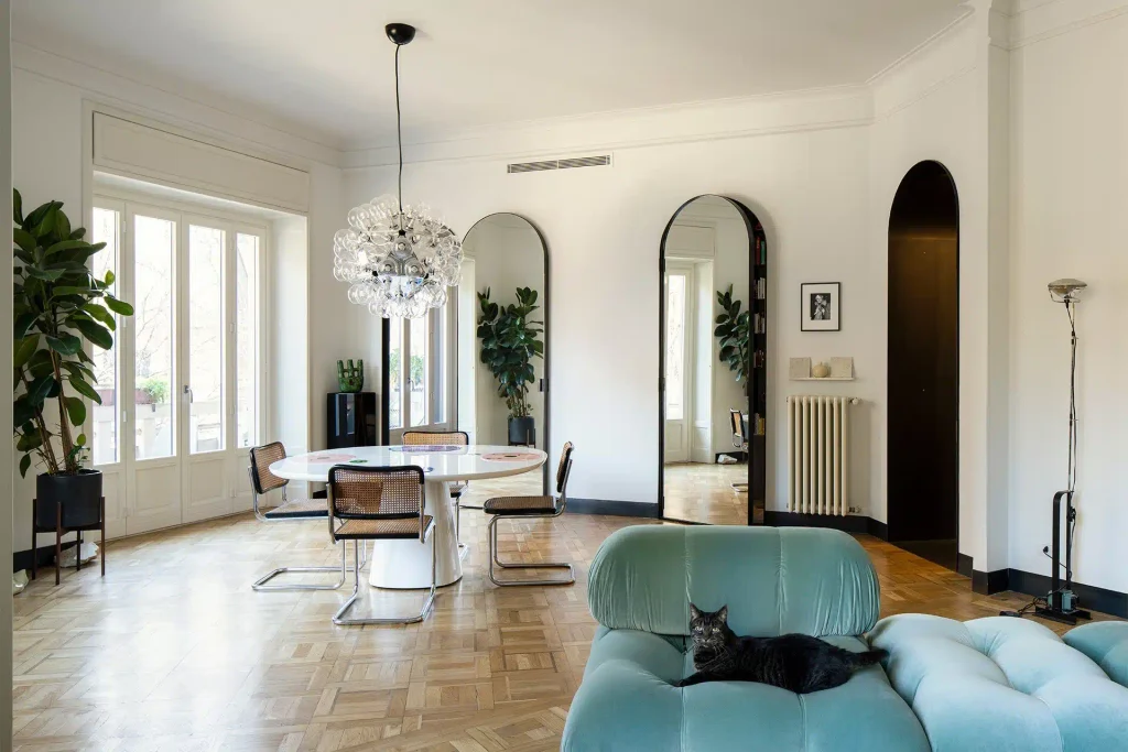 Bright modern living space featuring a black cat resting on a plush teal armchair, a white round dining table, and arched floor-length mirrors. top interior design firms in milan