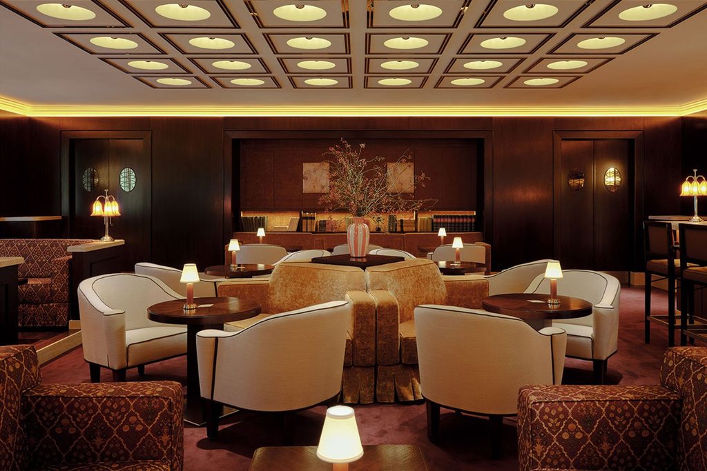 Dimly lit vintage-style lounge featuring dark wood wall paneling, coffered ceiling lighting, and cozy white armchairs. top interior design firms in milan