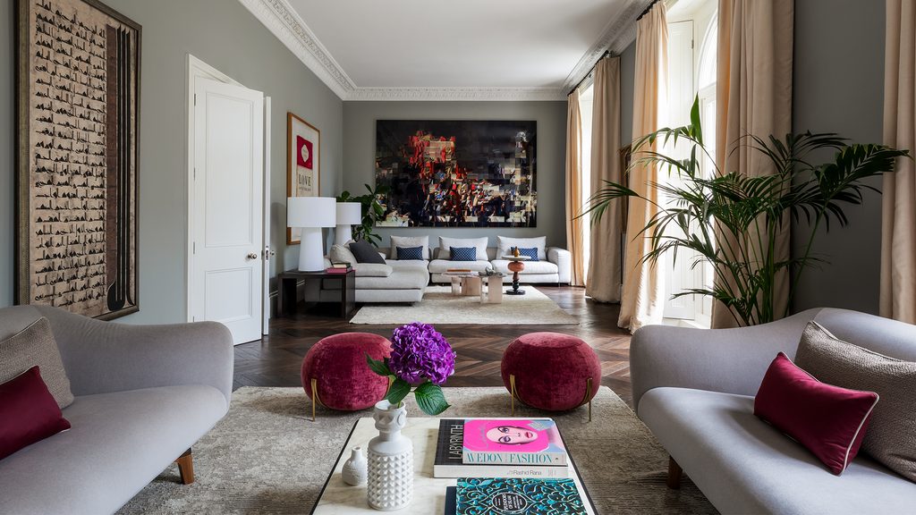 Elegant living room with tall windows, light grey sofas, dark wood floors, and rich berry-red poufs and accent pillows. top interior design firms in milan
