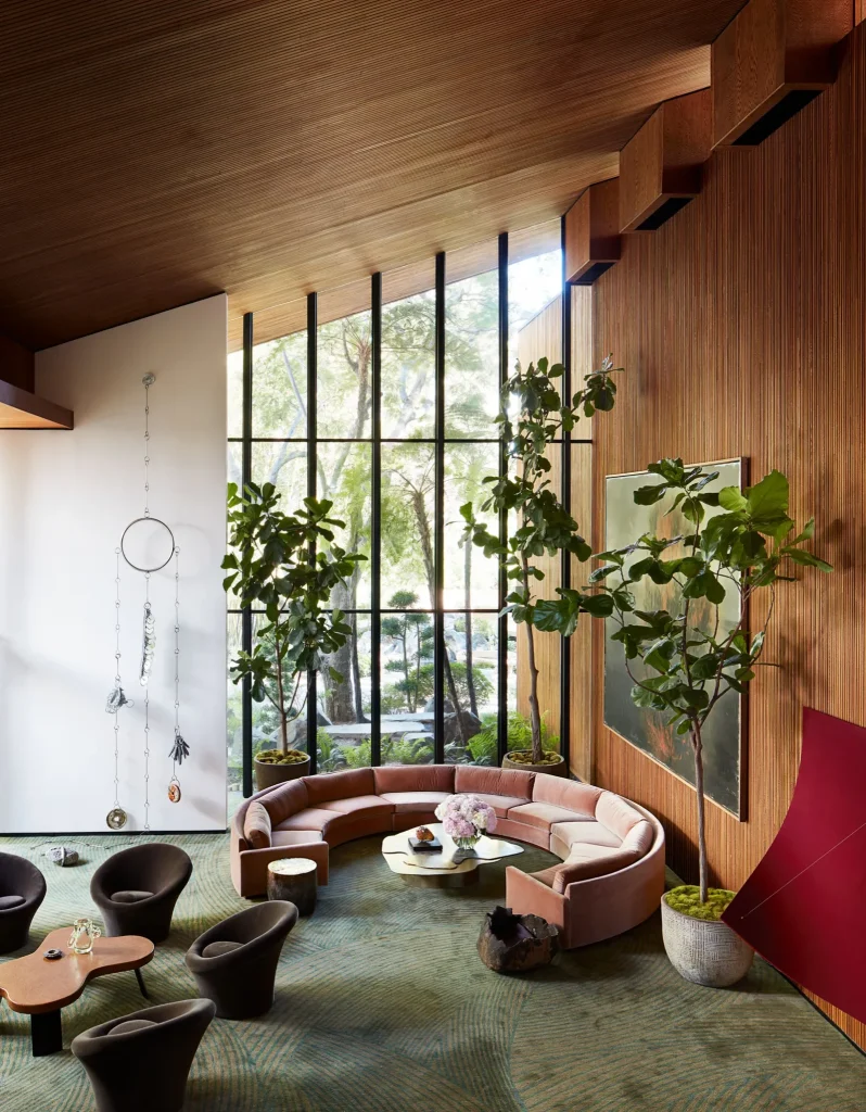 Spacious room with high ceilings, large glass windows, a curved light-pink sofa, a green rug, and wood-paneled walls.